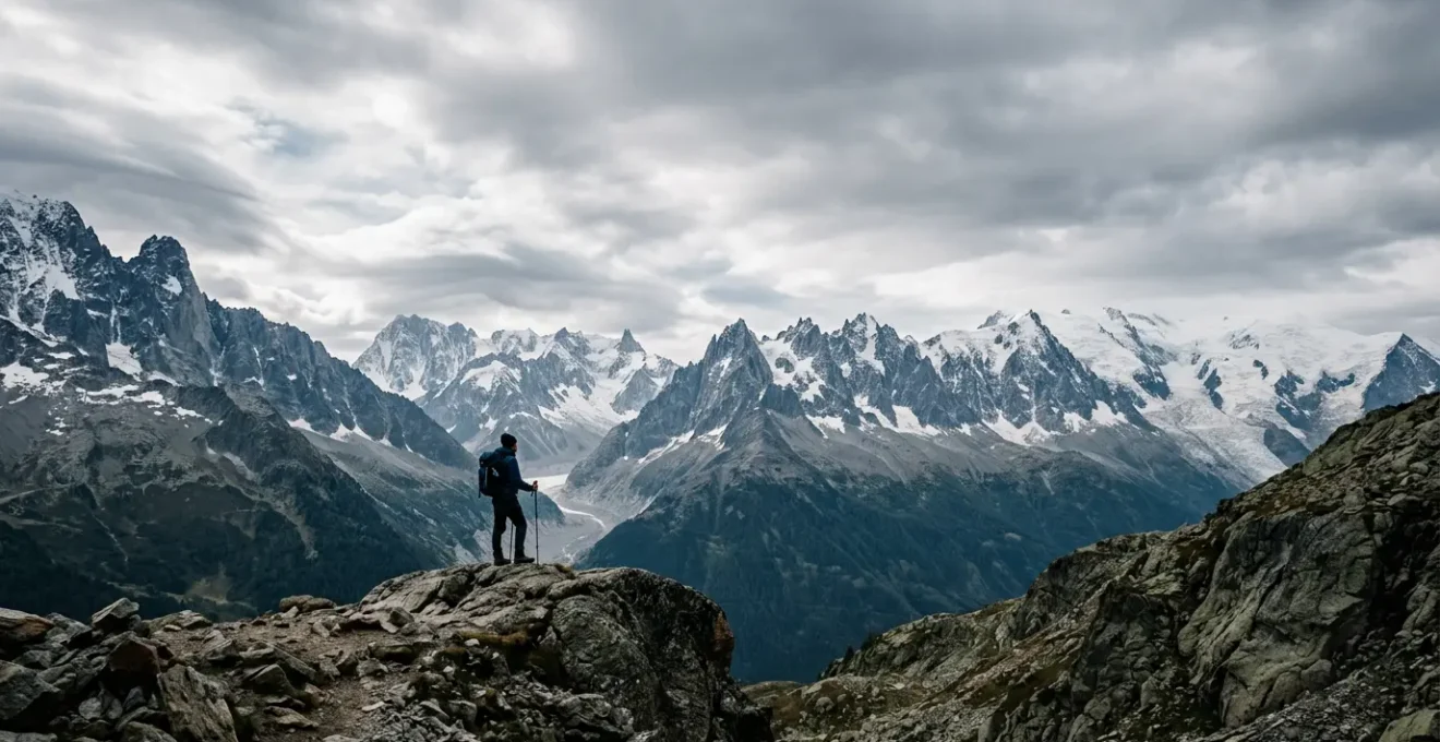 Mountaineer standing alone on rocky outcrop gazing toward distant snow-capped peaks under dramatic sky